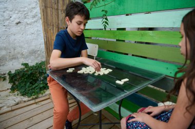 Cam bir masada domino oynayan iki çocuk, gündelik ortamda eğlenceli ve eğitici bir oyun oynuyorlar..