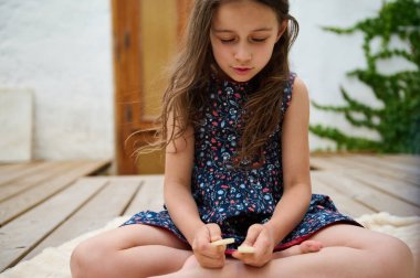 Çiçekli elbiseli genç bir kız ahşap bir güvertede oturuyor, sopalarla oynuyor. Etrafı yeşilliklerle çevrili, huzurlu bir anın tadını çıkarıyor..