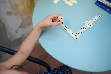 Bir çocuğun eli domino taşını tutuyor, açık havada mavi bir masada oyun oynuyor. Sahne boş zamanları yakalar, stratejiyi yakalar ve eğlenceli bir ortama odaklanır..
