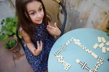 Genç bir kız açık hava masasında büyük bir hevesle domino oynar. Yüzündeki ifade neşe ve merakı gösteriyor. Bir anlık eğlence ve öğrenme anı yakalıyor..