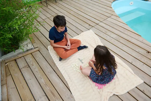 stock image Two kids play with word tiles on a blanket by the pool. The setting evokes summer fun, relaxation, and youthful creativity.