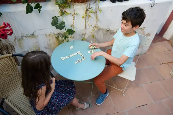 Bir kız ve erkek bahçede domino oynuyor, bitkilerle çevrili eğlenceli ve rahat bir oyunun tadını çıkarıyorlar..