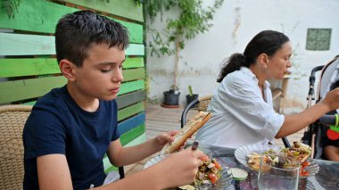 Bir aile, hoş bir bahçe ortamında çeşitli yemekler sunan açık havada rahat bir yemeğin tadını çıkarır. Beraberliğin ve sıradan yemeklerin özünü yakalar..