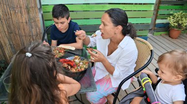 Bir aile bir masanın etrafında toplanmış güvertede yemeğin tadını çıkarıyordu. Açık hava manzarası ve canlı çitler bu birliktelik ve neşe anına sıcaklık katar..