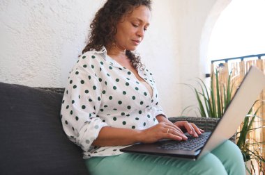 Polka noktalı tişörtlü bir kadın kanepe üzerinde dinlenirken dizüstü bilgisayar kullanıyor, bitkilerle çevrili, uzaktan kumandalı bir işi veya gelişigüzel ayarları vurguluyor..