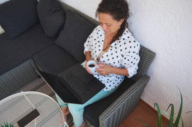 Bir kadın rahat bir terasta dizüstü bilgisayarıyla çalışırken kahveden zevk alır. Rastgele ve rahat ortam modern iş-yaşam dengesini gösterir.