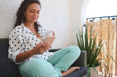 Bir kadının rahat bir balkonda akıllı telefonunu karıştırırken bir fincan kahvenin keyfini çıkardığı huzurlu bir sahne..