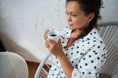 Beyaz benekli tişörtlü bir kadın rahat bir sandalyede otururken kahvenin tadını çıkarıyor. Huzurlu bir rahatlama ve tefekkür anı.