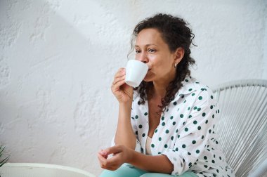 Bir kadın rahat ve aydınlık bir ortamda huzur içinde kahveyi yudumluyor, rahatlama ve sükunet anı yakalıyor..