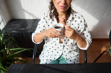 Puantiyeli tişörtlü bir kadın güneşli bir odada kahve fincanı tutarak rahatlama ve rahatlama anı yakalıyor..