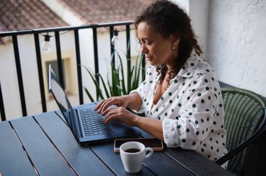 Bir kadın rahat bir balkonda çalışırken dizüstü bilgisayarına odaklanır. Akıllı telefonu ve kahvesi olan sıradan bir iş ortamından hoşlanıyor..