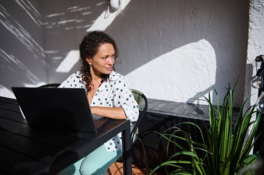 Güneşli açık havada dizüstü bilgisayarla uzaktan çalışan kendine güvenen bir kadın, görevlere odaklanıyor. İş-yaşam dengesini, doğal ışığı ve üretkenliği kucaklamak.