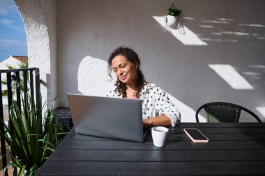 Bir kadın terasta uzaktan çalışmayı sever. Yanında bir fincan kahve ve akıllı telefonla bilgisayarını kullanır. Açık hava verimliliğini kucaklar..