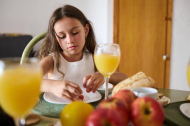 Genç bir kız, taze meyve, portakal suyu ve sıcak bir yemek masasındaki ekmekle çevrili kahvaltısına odaklanıyor..