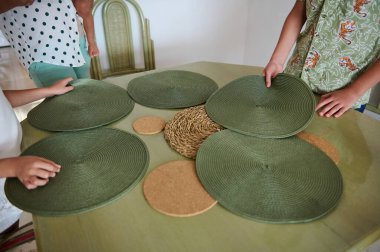 Family members are setting a table with green placemats and woven decor, highlighting a casual and warm atmosphere for a gathering. clipart