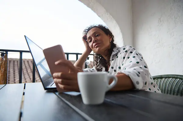 stock image A relaxed woman in a polka dot shirt interacts with a smartphone and laptop on an outdoor patio table, accompanied by a coffee cup.