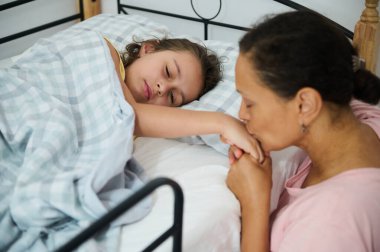 Anne sevgi dolu bir şekilde kızının elini öpüyor. Yatakta yatarken şefkati ve şefkati simgeliyor. Aile sıcaklığını yakalar ve rahatlatır..