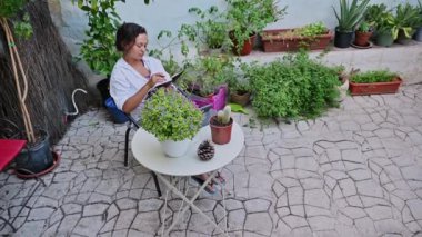 Bir kadın verandada huzurlu bir andan zevk alır, bir deftere yazarak, yemyeşil saksılarla çevrili..