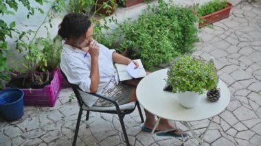 Bir kadın düşünceli bir şekilde açık bir masada oturur ve defterine bir şeyler yazar. Çevresi canlı yeşil bitkilerle çevrili. Huzurlu ve yaratıcı bir atmosfer yaratıyor..