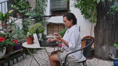 Sakin bir bahçede dizüstü bilgisayarı ve telefonu olan bir kadın doğal ofis ortamında uzaktan çalışma kavramını vurguluyor..