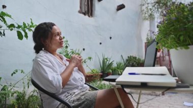 Dışarıda dizüstü bilgisayarıyla oturan bir kadın uzaktan çalışma toplantısına katılıyor. Çevresi yeşillik ve doğal elementlerle çevrili. Sakin ve odaklanmış bir ortam yaratıyor..