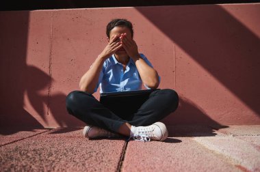 Mavi gömlekli bir kişi dışarıda dizüstü bilgisayarla bağdaş kurarak oturur, yüzünü hayal kırıklığı ya da gün ışığı altında stres ile kapatır..