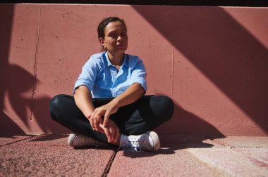 Güneşin altında bağdaş kurmuş, pembe bir duvara yaslanmış kentsel bir ortamda huzurlu ve rahat bir anı yakalayan bir insan..