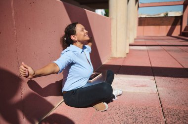 Neşeli bir kadın açık bir koridorda oturuyor, gözleri kapalı ve gülümsüyor. Huzuru ve rahatlığı kucaklayarak, güneşin sıcaklığından ve rutinden kısa bir kaçıştan zevk alır..