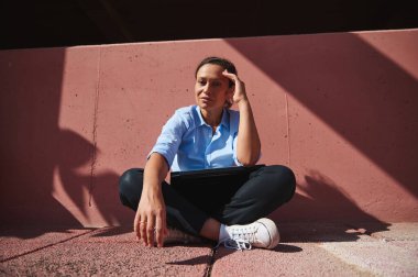 Dışarıda kucağında dizüstü bilgisayarla oturan bir kadın için yansıtıcı bir an. Güneş ışığında sırılsıklam, düşüncelere dalmış, arkasında pembe beton bir duvarla, derin düşüncelere dalmış..