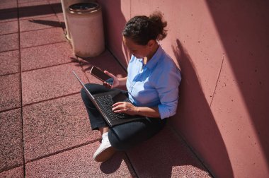 Dizüstü bilgisayarı ve akıllı telefonu olan bir kadın pembe çatı terasında oturmuş güneşli günün tadını çıkarıyor..