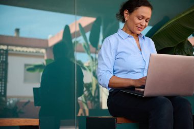 Dışarıda dizüstü bilgisayarıyla çalışan gülümseyen bir kadın, verimliliği ve dinginliği rahat bir ortamda yansıtıyor. Cam yansıması dışarıdaki iş yerine benzersiz bir boyut ekler..