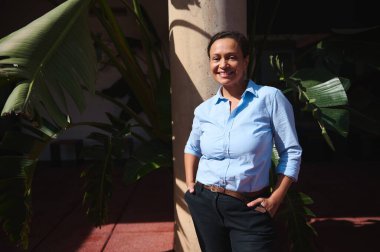 Kendine güvenen bir kadın dışarıda duruyor, yumuşak güneş ışığında sıcak bir şekilde gülümsüyor çevrende yeşil tropikal bitkiler, pozitiflik ve doğayı aktarıyor..