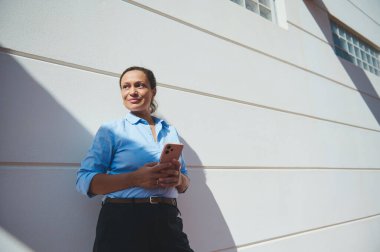 Mavi gömlekli profesyonel bir kadın telefonunu açık havada tutuyor. Güneş ışığı modern bir binaya gölge düşürüyor..