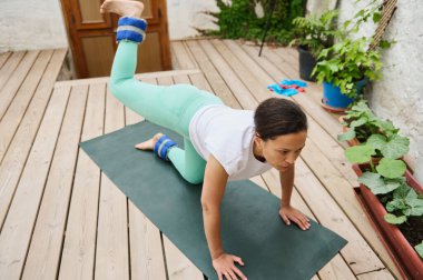 Bir kadın ahşap bir güvertede yoga yapıyor, bilek ağırlıklarını kullanarak dengeyi ve gücü arttırıyor, yemyeşil bitkilerle çevrili..
