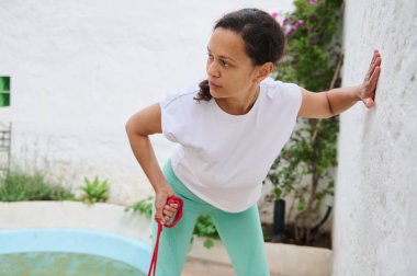Kadın bir bahçede direnç bandıyla spor yapıyor. Spor kıyafetleri giyerken güç ve dengeye odaklanıyor..