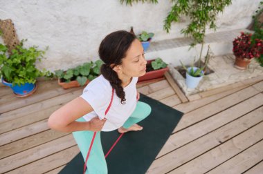 Tahta bir güvertede yoga yapan bir kadın, yemyeşil bitkilerle çevrili, açık hava ortamında rahatlama ve farkındalığı teşvik ediyor..