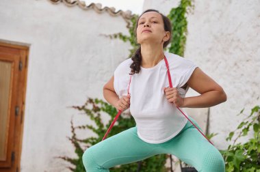Kararlı bir kadın dışarıda bir direniş bandıyla egzersiz yapıyor. Spor salonunun güneşli ve huzurlu bahçesine odaklanıyor..