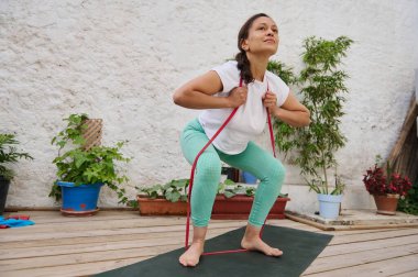 Bir kadın yoga minderinin üzerine çömelmiş, dingin bir bahçe ortamında bitkilerle çevrili, fitness ve sağlıklı yaşam tarzını destekleyen bir direniş bandına bağlanır..