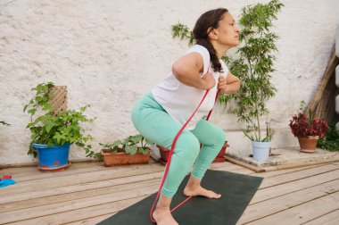 Bir kadın, dışarıda tahta bir güvertede direniş bandı egzersizi yapıyor. Etrafı saksı bitkileriyle çevrili fitness ve dayanıklılık eğitimine odaklanmış..