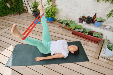 Bir kadın, bahçedeki yoga minderinde direniş bandı kullanarak egzersiz yapıyor. Ayarda saksı bitkileri ve ahşap bir güverte yer alıyor..