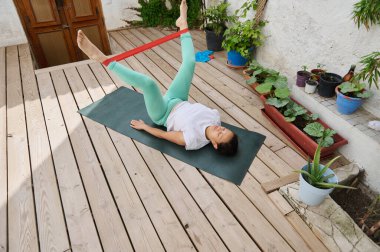 Bir kadın, güneş yanığı ahşap bir güvertede direnç bandı kullanarak yoga yapıyor. Titreyen saksı bitkilerinin ortasındaki zindelik ve gevşemeyi arttırıyor..