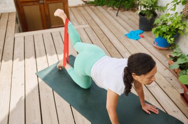 Güvertede direniş bandıyla yoga yapan odaklanmış bir kadın esnekliği ve gücü arttırıyor. Açık bir alanda sakinlik ve odaklanma.