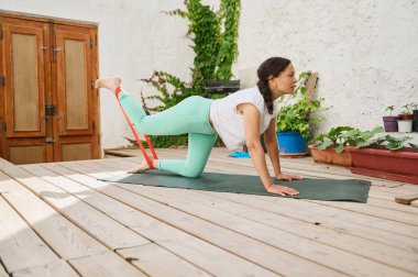Bir kadın ahşap bir güvertede egzersiz yaparken direnç bandı kullanıyor, spor yapmayı ve açık havada sağlıklı bir yaşam tarzını teşvik ediyor..