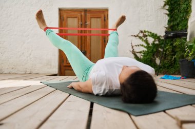 Dışarıdaki sakin bir bahçede yoga minderinde egzersiz yaparken direniş bandı kullanan biri. Sağlık ve zindeliğe odaklan.