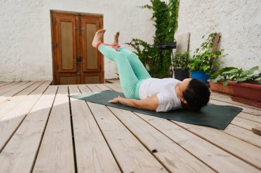 Dışarıdaki ahşap güvertede yoga egzersizleri yapan bir kadın. Etrafı bitkilerle çevrili. Sahne sakin ve odaklanmış bir atmosfer yakalar..
