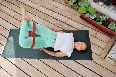 Bir kadın, açık havada yoga minderinde direnç bantlarıyla pilates egzersizleri yapıyor. Sakin bir bahçe ortamında spor ve esnekliği vurguluyor..