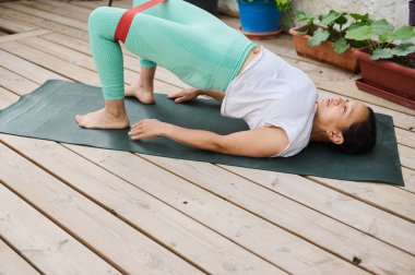 Tahta bir güvertede yoga yapan bir kadın, dayanıklılığını, esnekliğini ve dengesini arttırmak için köprüdeki direnç bandını kullanır..