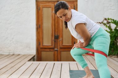 Bir kadın dışarıda tahta bir güvertede direnç bandı egzersizi yapıyor, dayanıklılık gösteriyor ve günlük kıyafetlere odaklanıyor. Açık hava fitness ve refah konsepti.