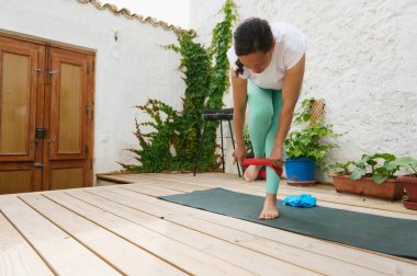 Kadın, açık hava güvertesinde bir direniş bandıyla yoga yapıyor, dengeyi ve dengeyi bir bahçe ortamında somutlaştırıyor..