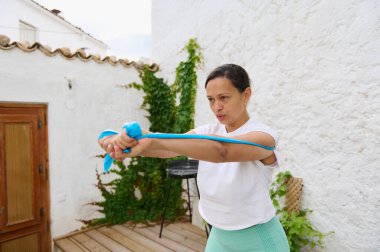 Odaklanmış bir kadın açık terasta direnç bandı çalışması yapıyor. Doğal ortamdaki güç ve uygunluğu vurguluyor..